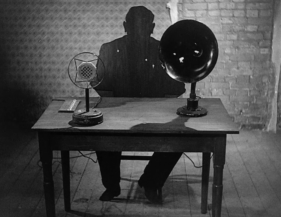 Film still from The Testament of Dr. Mabuse: Mabuse's shadow looms behind a desk with a microphone and speaker horn.