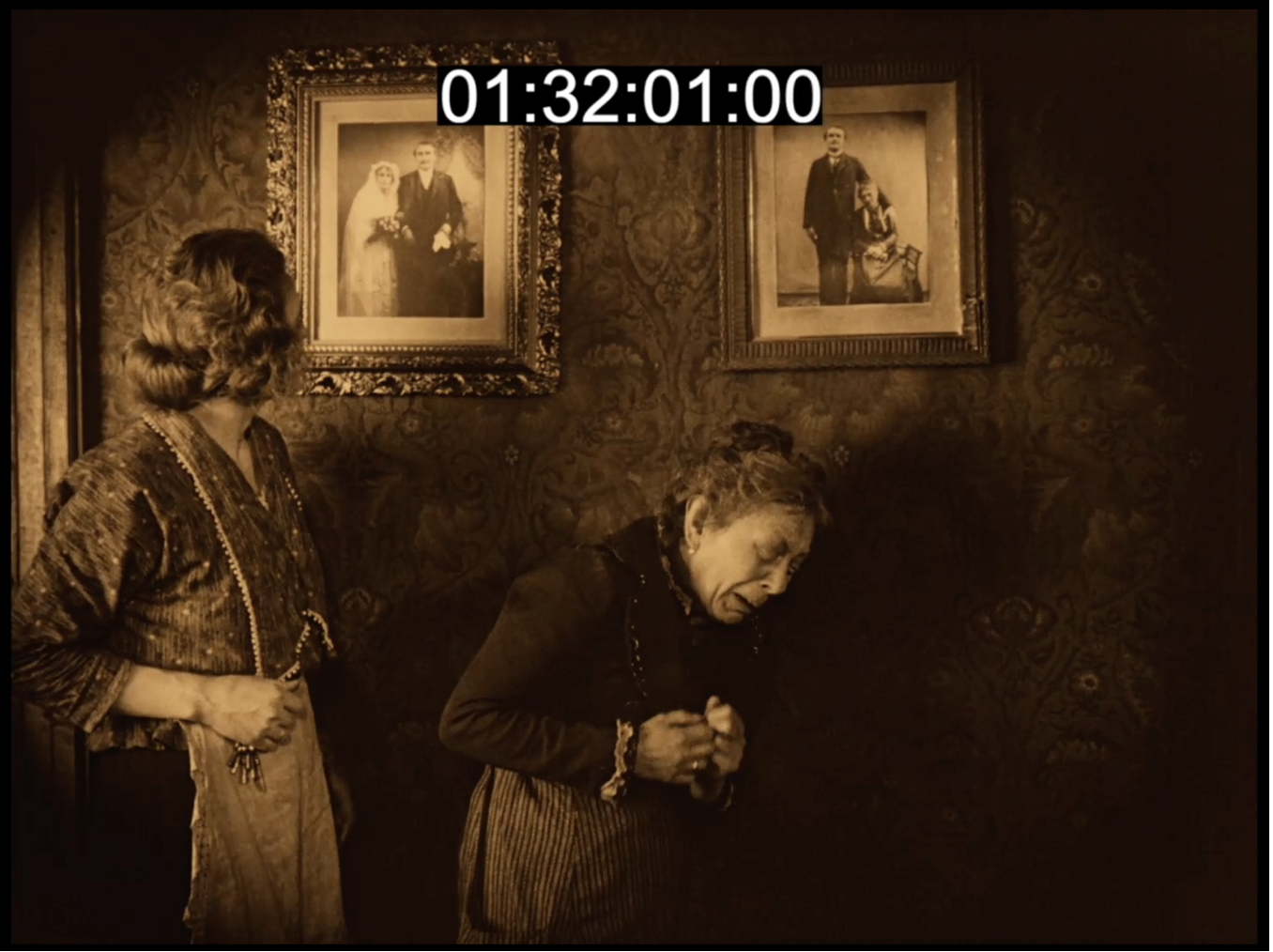 Film still from Sylvester: two women stand beneath framed family wedding portraits on patterned wallpaper.