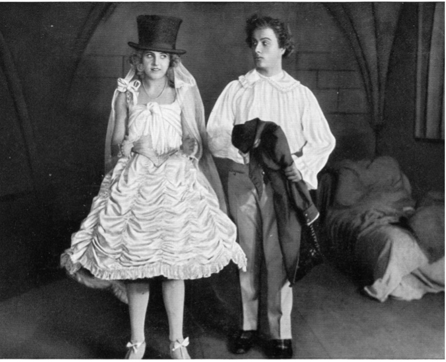 Film still: a figure in a wedding dress and top hat stands beside a young man in a vaulted stone room.