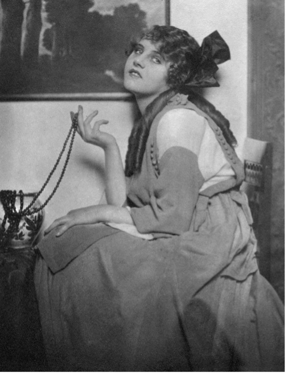 Portrait of Ossi Oswalda seated, holding a string of beads, gazing upward.