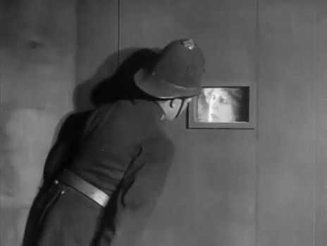Film still: a police officer in a domed helmet peers through a small hatch in a cell door at a woman's face inside.