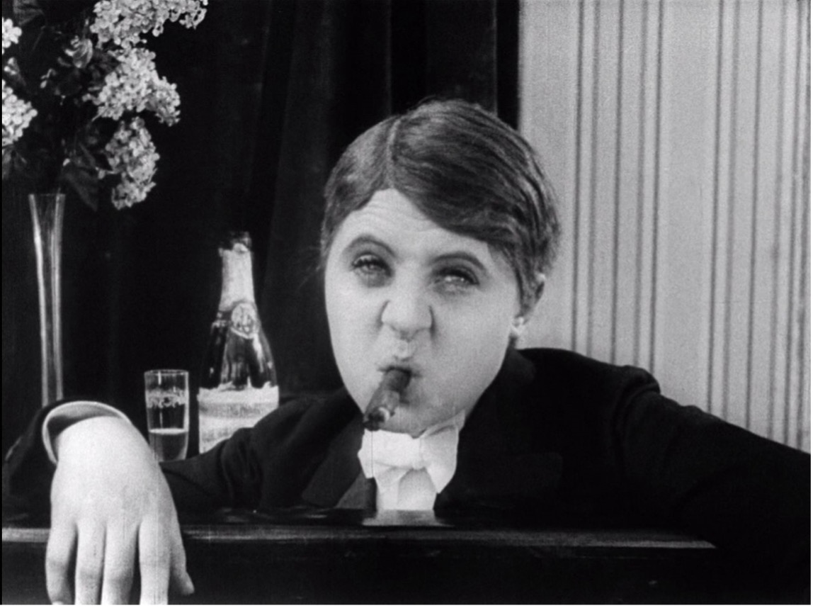 Film still from Ich möchte kein Mann sein (I Don't Want to Be a Man): Ossi Oswalda in male evening dress, smoking a cigar beside a champagne bottle.