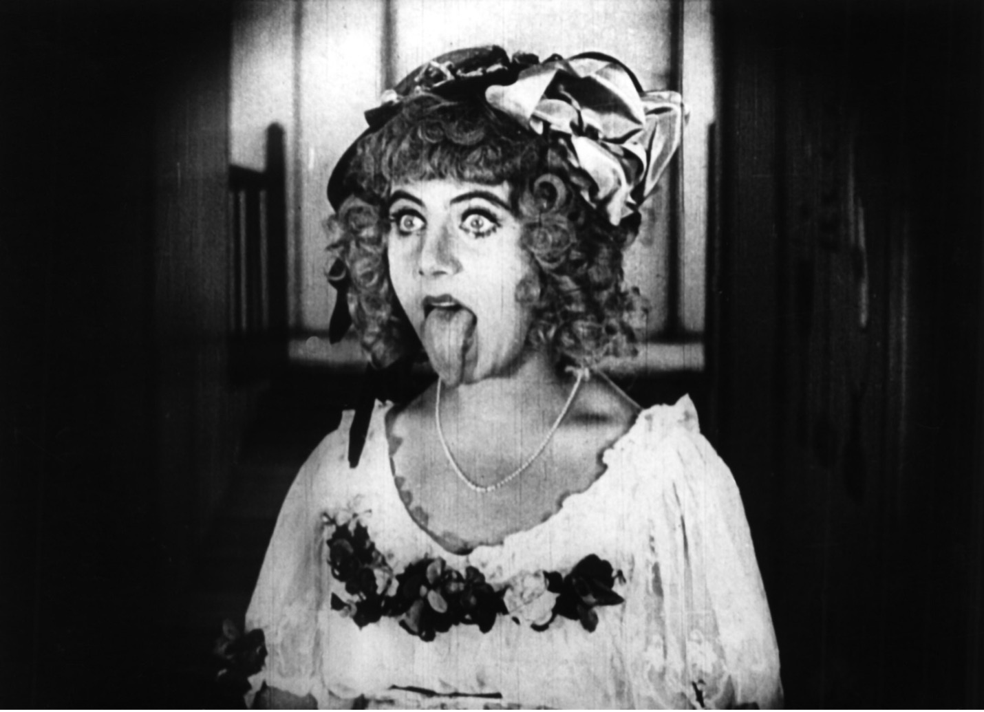 Film still: a performer in a bonnet with a large bow sticks out her tongue in an exaggerated grimace.