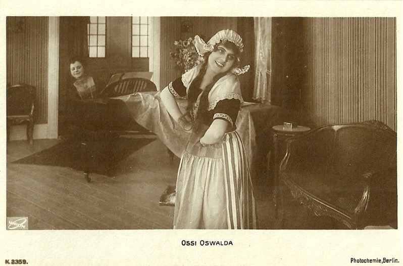 Postcard portrait of Ossi Oswalda smiling in costume, with a man seated at a piano in the background.