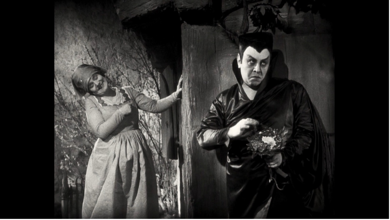 Film still: a devil-costumed figure with dark eye makeup and small horns stands beside a woman in peasant dress in a doorway.