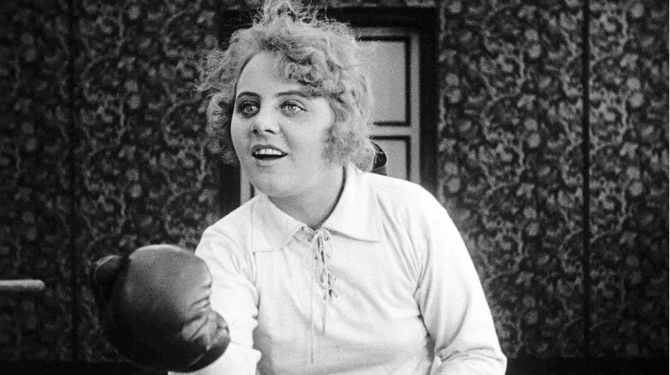 Film still: Ossi Oswalda, smiling, throws a punch wearing a boxing glove.