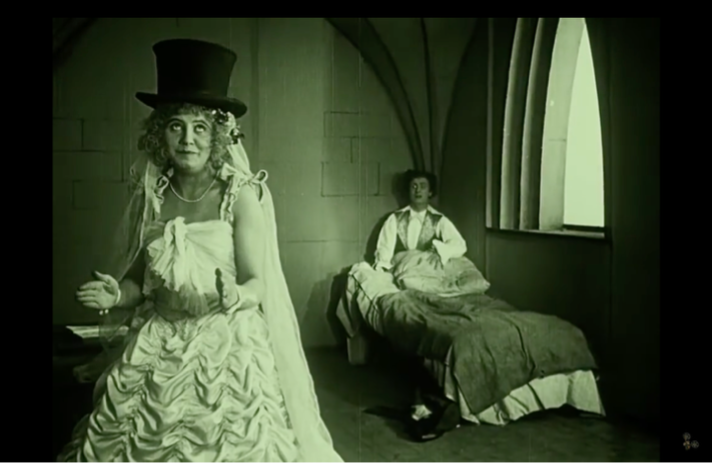 Film still from Die Puppe: a figure in a wedding gown and top hat stands in a vaulted stone room beside another figure seated on a bed.