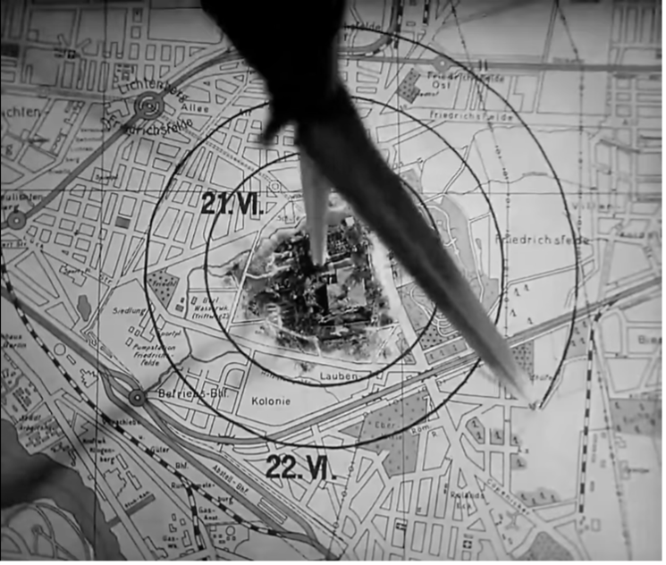 Film still from M: a near-overhead view of the compass, its top blurred, tracing a circle on the map around the miniature garden house.