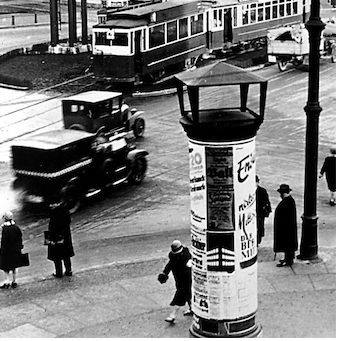 Elevated view of a Berlin street: a poster-covered Litfaßsäule in the foreground, a tram and a motor car crossing behind it, pedestrians on the pavement